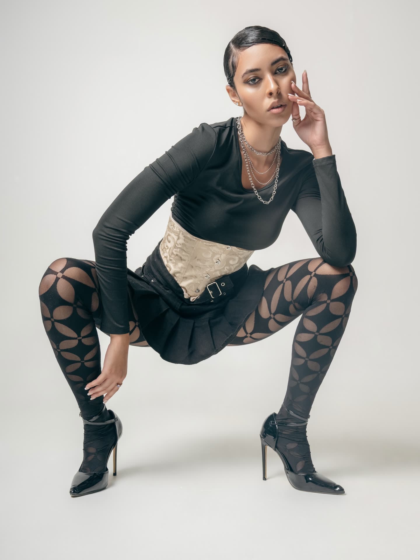 Jess crouched wide in a black long-sleeve top, cream brocade corset belt and floral cut-out tights with stiletto boots