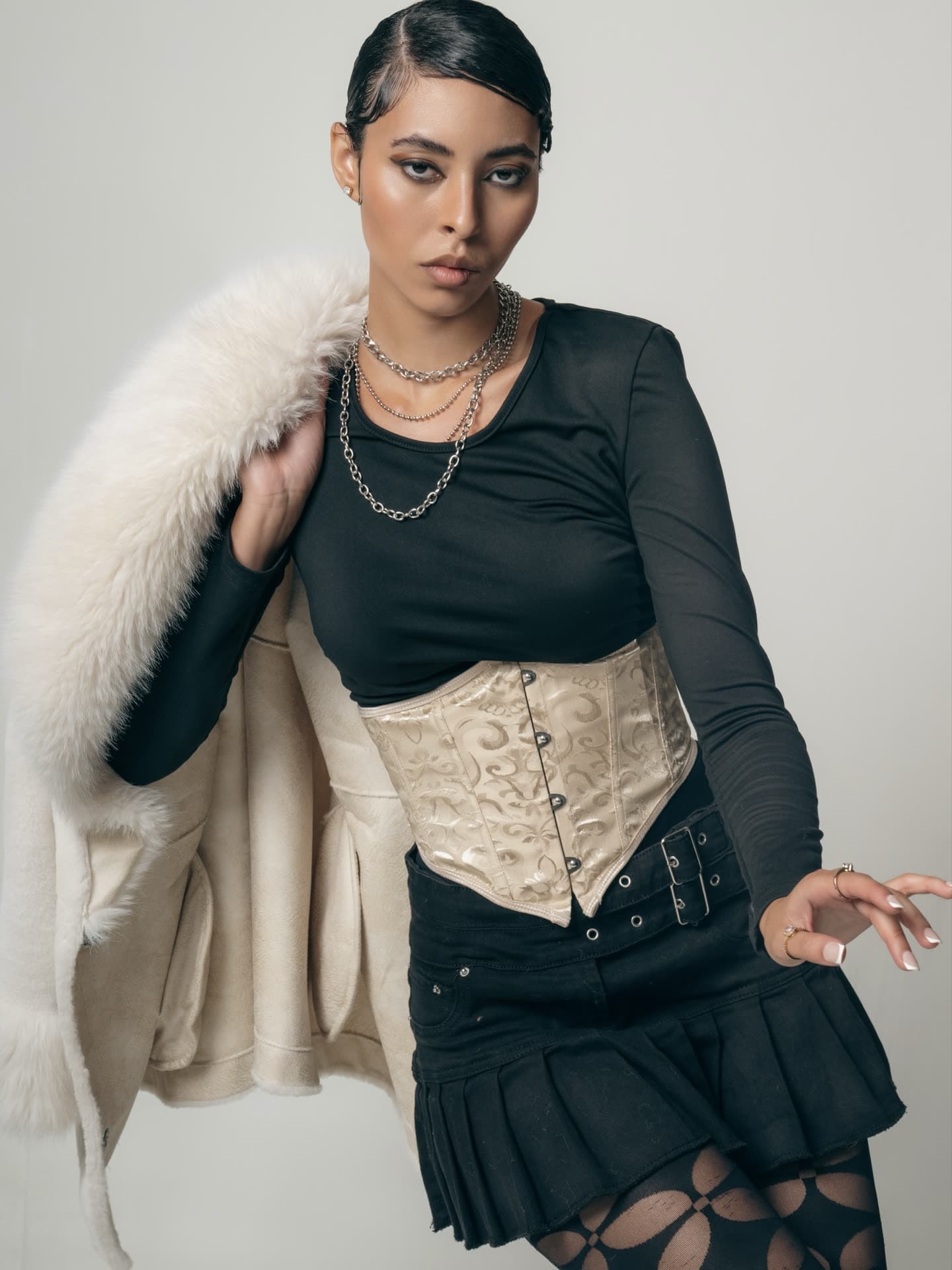 Jess with a white fur stole over one shoulder, cream corset and pleated black mini skirt, hand raised