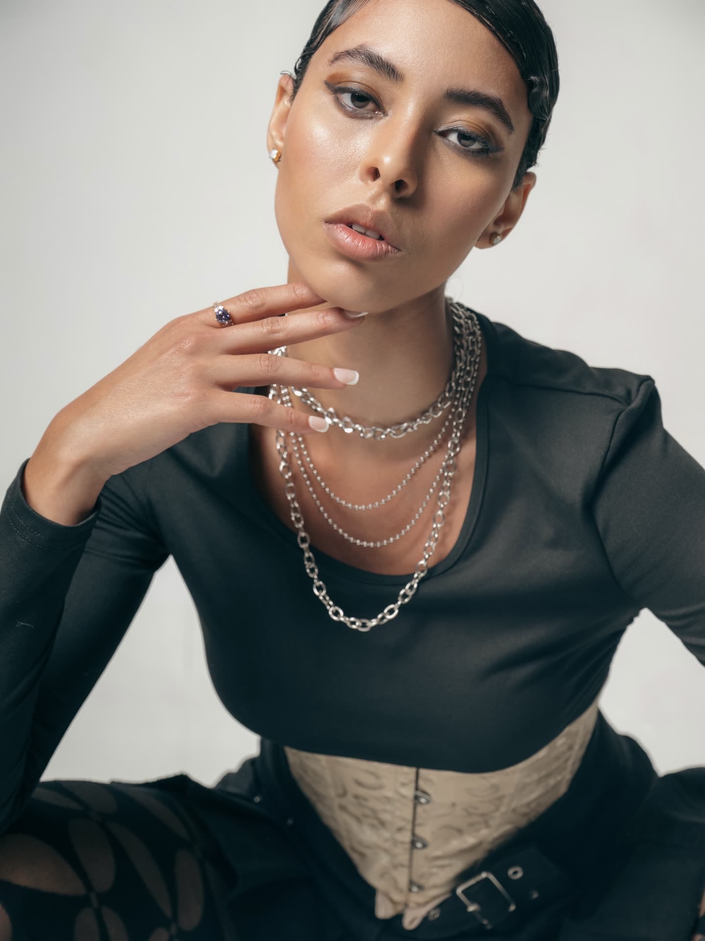 Close beauty portrait, hand resting under her chin, layered silver chains and slicked-back hair