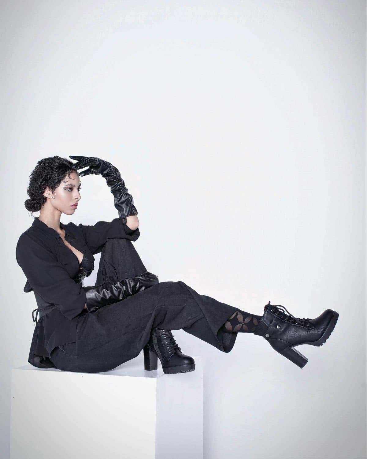 Jess in head-to-toe black tailoring, seated on a white plinth, braids raised overhead