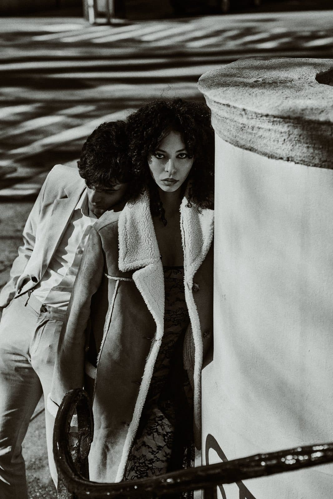 Jess and a fellow model in an oversized trench and shearling coat, black and white editorial, leaning against a weathered column