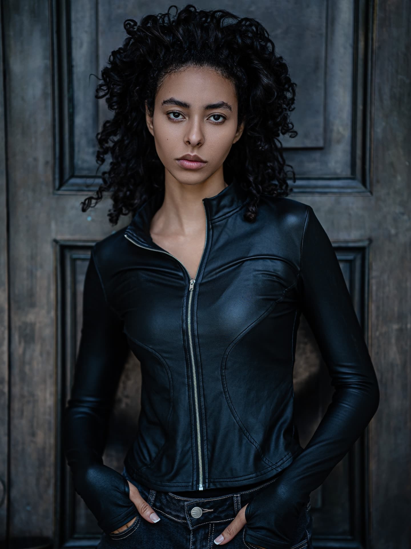 Jess in a fitted black leather moto jacket and dark jeans, hands in her pockets, against a weathered blue-grey door