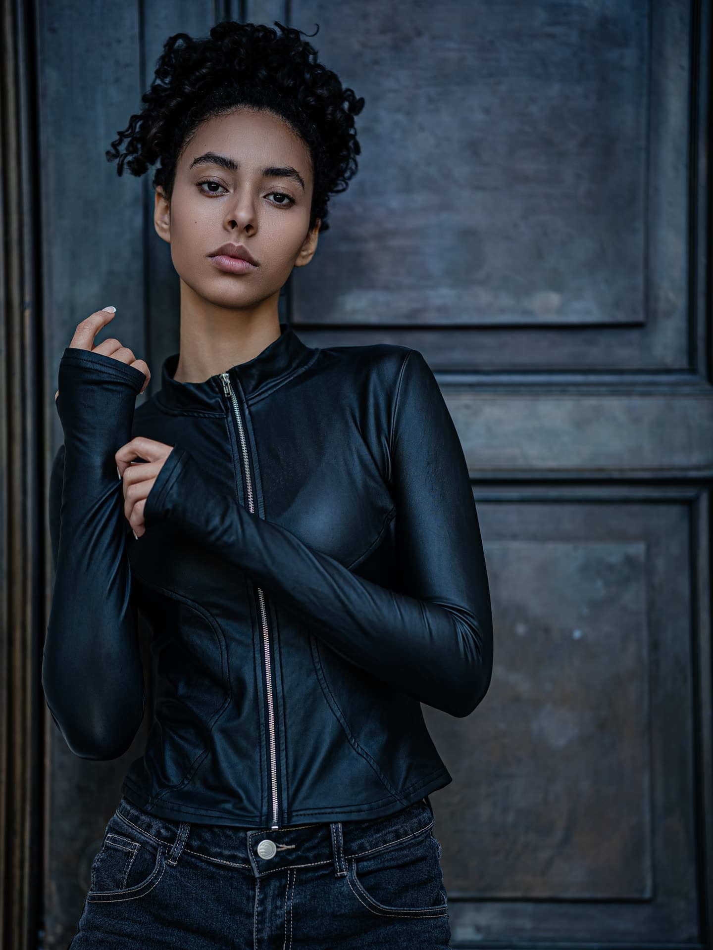 Jess in the black leather jacket with hands raised to her chest, curls loose, weathered door behind
