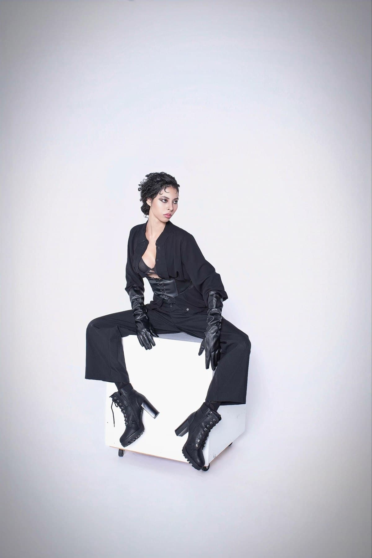 Jess seated in an all-black look with long leather gloves and lace-up boots, studio portrait