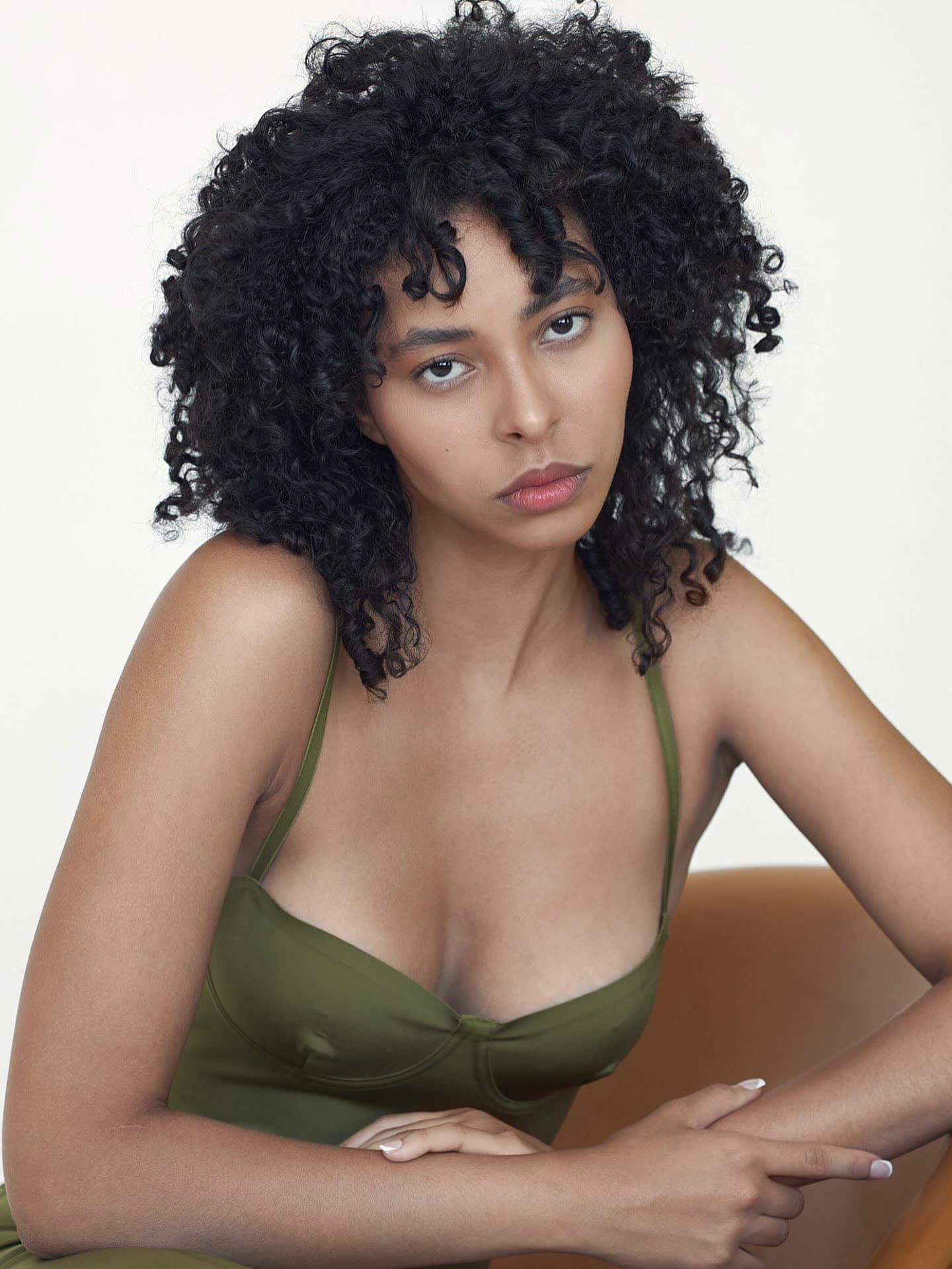 Jess leaning forward on the tan chair with arms crossed, olive dress, sage backdrop