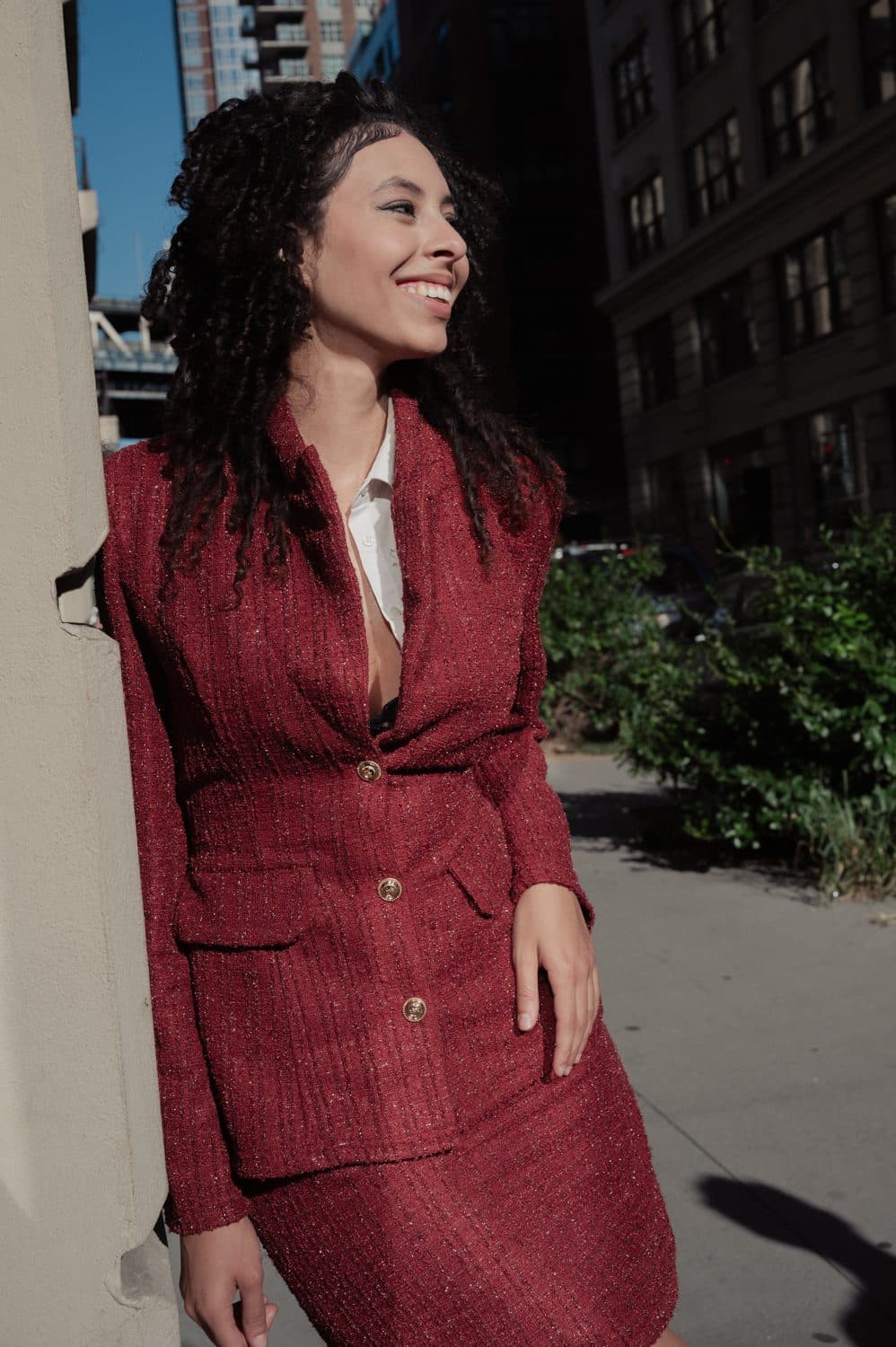 Jess in a scarlet tweed skirt suit on a sunlit sidewalk, looking over her shoulder